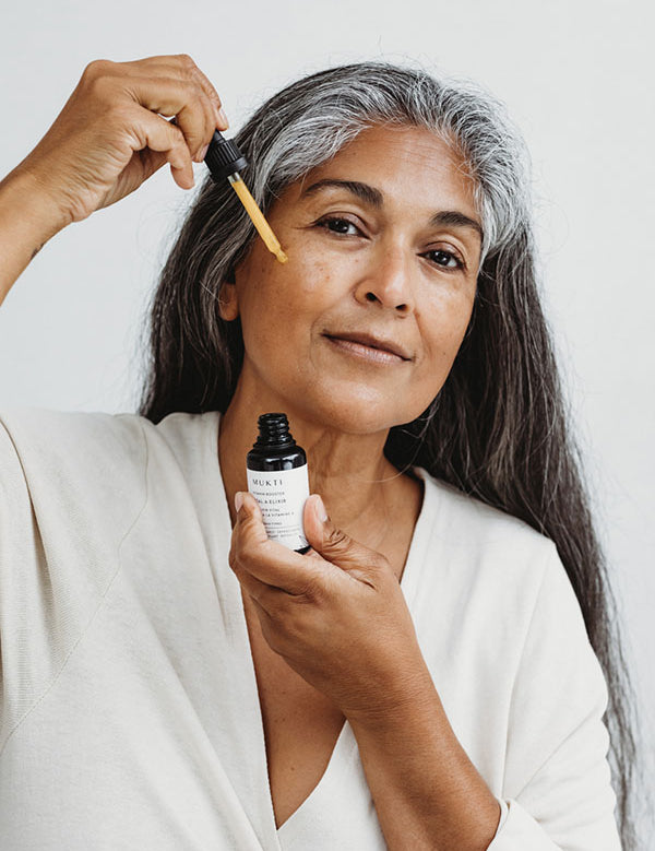 Woman with long grey hair applying Mukti Vital A Vitamin Booster to her face with the dropper
