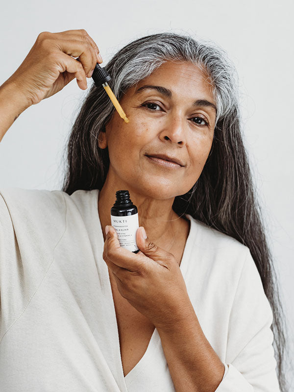 Woman with long grey hair applying Mukti Vital A Vitamin Booster to her face with the dropper