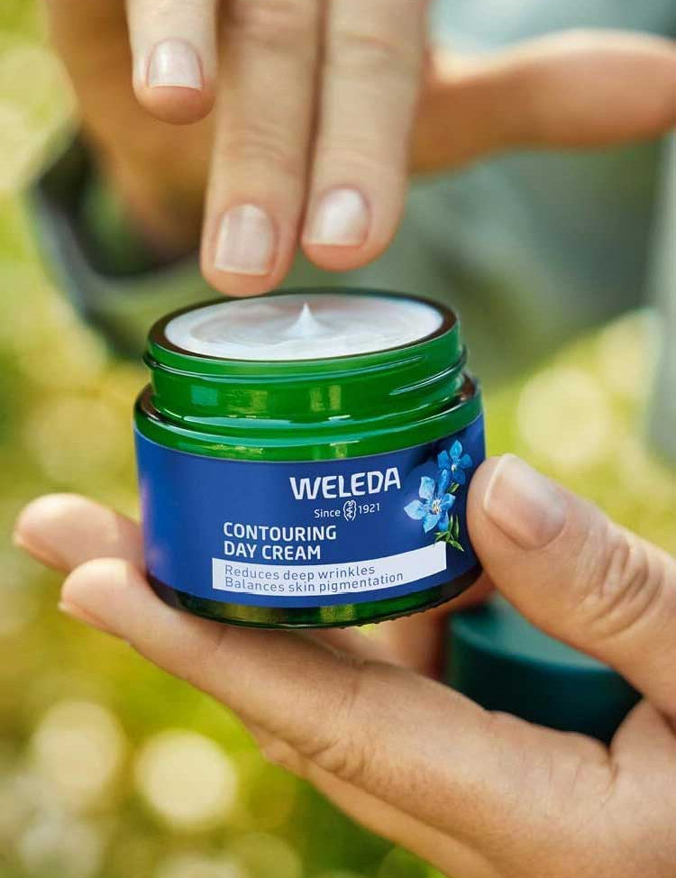 A person holds an open jar of Weleda Contouring Day Cream Blue Gentian & Edelweiss, lifting the lid and dipping a finger into the cream. The green jar with a blue label is shown outdoors in natural light.