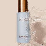 Pale blue INIKA Hyaluronic Hydration Complex bottle with a gold cap, surrounded on the right and below by a large splash of clear water against a beige background.