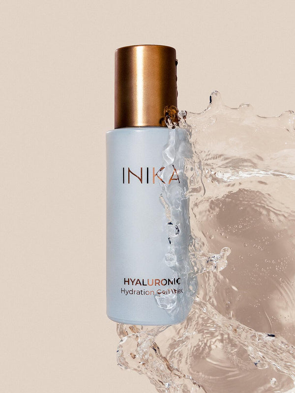 Pale blue INIKA Hyaluronic Hydration Complex bottle with a gold cap, surrounded on the right and below by a large splash of clear water against a beige background.