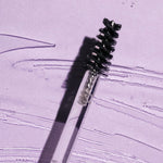 Black spiral brow brush on a clear wand lying diagonally across a lilac surface streaked with clear gel, casting a sharp shadow.
