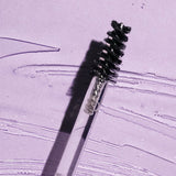 Black spiral brow brush on a clear wand lying diagonally across a lilac surface streaked with clear gel, casting a sharp shadow.
