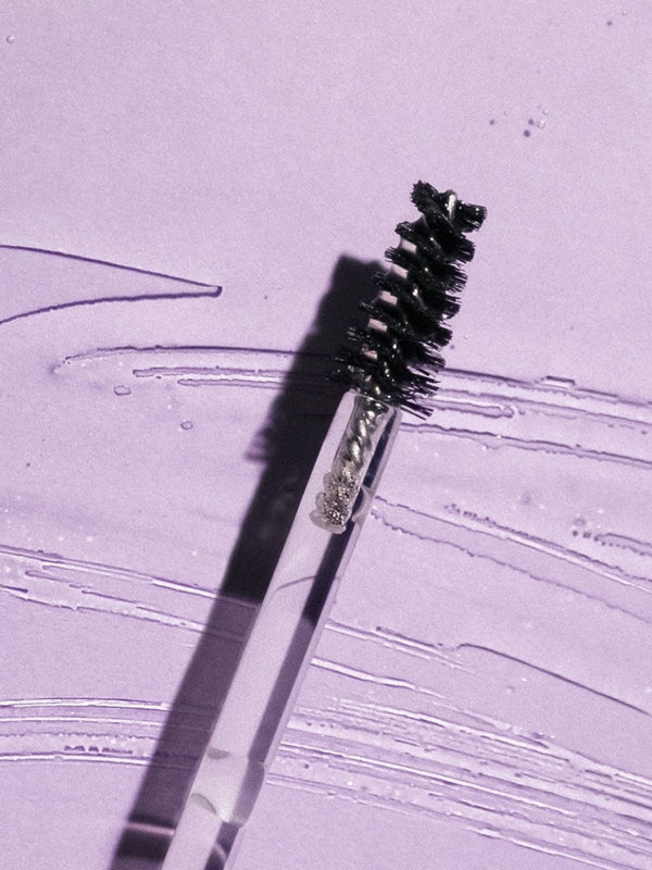 Black spiral brow brush on a clear wand lying diagonally across a lilac surface streaked with clear gel, casting a sharp shadow.