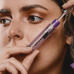 Woman holding an open clear-and-purple Madara Grow Fix booster tube diagonally beside her face, with the wand lifted from the tube above her cheek.
