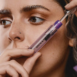 Woman holding an open clear-and-purple Madara Grow Fix booster tube diagonally beside her face, with the wand lifted from the tube above her cheek.