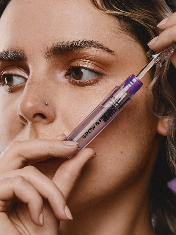 Woman holding an open clear-and-purple Madara Grow Fix booster tube diagonally beside her face, with the wand lifted from the tube above her cheek.