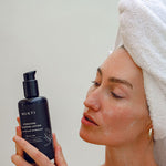 Woman in a towel turban reading the Mukti Hydrating Cleansing Lotion label