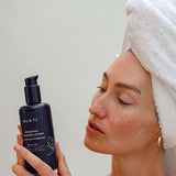 Woman in a towel turban reading the Mukti Hydrating Cleansing Lotion label