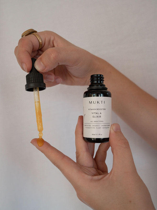 Hands drawing golden serum from the Mukti Vital A Vitamin Booster bottle with the dropper