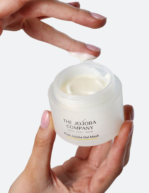 One hand holding an open jar of Pure Jojoba Gel Mask while a fingertip lifts a small dollop of its white gel cream.