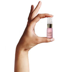 Hand holding a miniature lilac Neck & Decolletage Lifting Concentrate bottle between thumb and fingertips, showing its small size and gold pump under a clear cap.