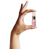 Hand holding a miniature lilac Neck & Decolletage Lifting Concentrate bottle between thumb and fingertips, showing its small size and gold pump under a clear cap.