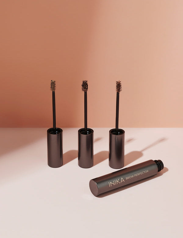 Three INIKA Brow Perfector wands with small brown-coated brushes stand upright in their dark metallic caps, with an open tube lying in front on a pink surface against a terracotta background.