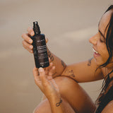 Smiling woman at the beach holding the Mukti Rose Blossom Hydrating Mist bottle