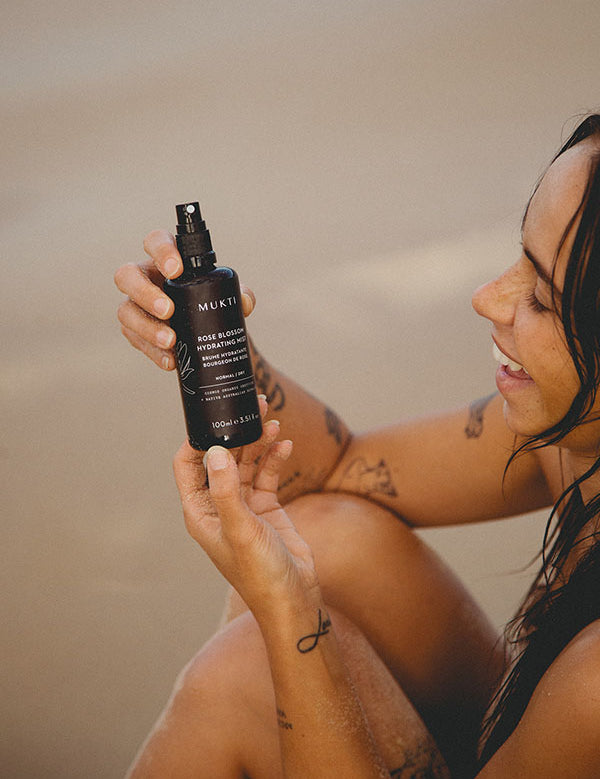 Smiling woman at the beach holding the Mukti Rose Blossom Hydrating Mist bottle