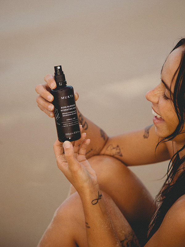 Smiling woman at the beach holding the Mukti Rose Blossom Hydrating Mist bottle