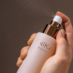 Hand holding a pale pink INIKA Hydrating Toning Mist bottle and pressing its white spray pump, releasing a fine mist into the air against a dark background.