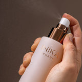 Hand holding a pale pink INIKA Hydrating Toning Mist bottle and pressing its white spray pump, releasing a fine mist into the air against a dark background.