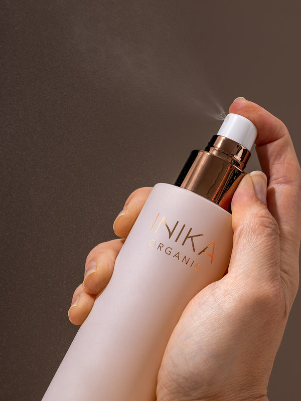 Hand holding a pale pink INIKA Hydrating Toning Mist bottle and pressing its white spray pump, releasing a fine mist into the air against a dark background.