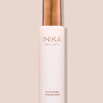 Tall pale pink INIKA Hydrating Toning Mist bottle with a cylindrical gold cap, standing upright and casting a narrow shadow on a beige background.