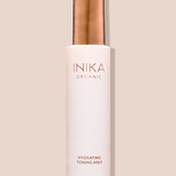 Tall pale pink INIKA Hydrating Toning Mist bottle with a cylindrical gold cap, standing upright and casting a narrow shadow on a beige background.