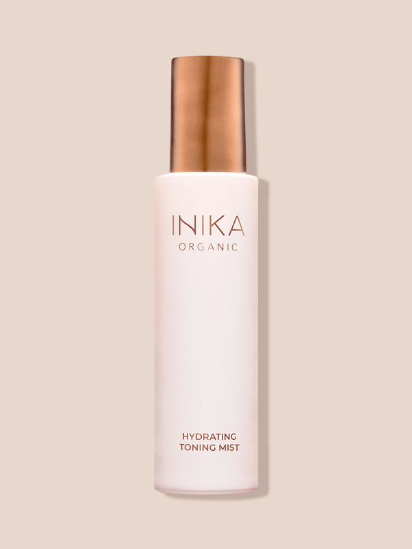 Tall pale pink INIKA Hydrating Toning Mist bottle with a cylindrical gold cap, standing upright and casting a narrow shadow on a beige background.