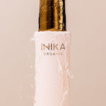 Close-up of water pouring onto the gold cap of a pale pink INIKA Hydrating Toning Mist bottle and running down its sides against a blush-pink background.