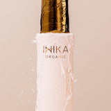 Close-up of water pouring onto the gold cap of a pale pink INIKA Hydrating Toning Mist bottle and running down its sides against a blush-pink background.