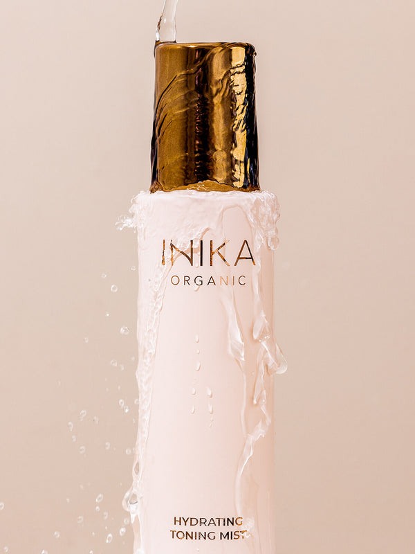Close-up of water pouring onto the gold cap of a pale pink INIKA Hydrating Toning Mist bottle and running down its sides against a blush-pink background.