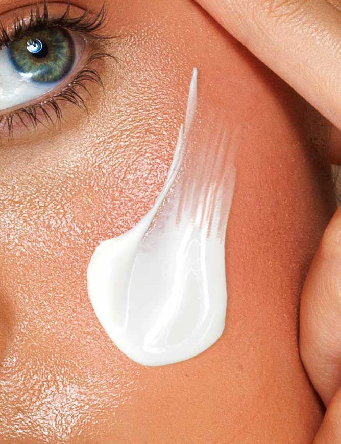 A close-up of a person’s smooth skin and blue eye, with a streak of Avocado Zinc SPF50 Natural Physical Face Sunscreen 100ml on their cheek as a finger gently touches the skin near the temple.