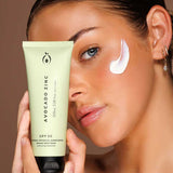 A woman holds a tube of Avocado Zinc SPF50 Natural Physical Face Sunscreen 100ml near her face, displaying a swipe of sunscreen on her cheek against a neutral background.
