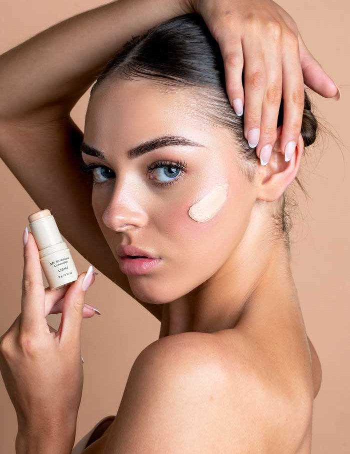 A blue-eyed woman with slicked-back hair holds Avocado Zinc SPF50 Natural Concealer - Light near her face, displaying a cream swatch on her cheek. She poses with one hand above her head against a peach background.