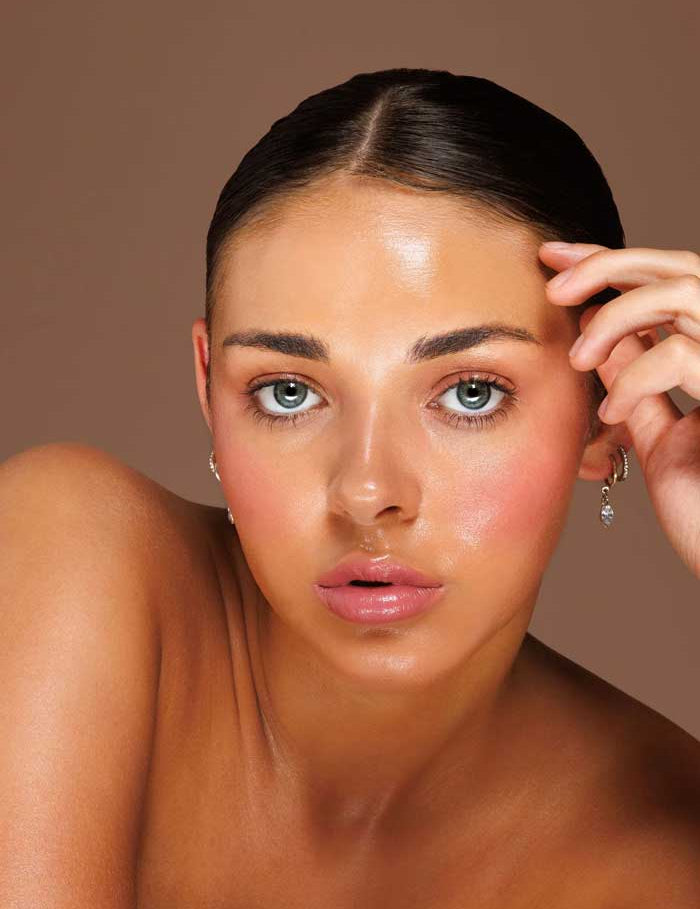 A woman with smooth, glowing skin and slicked-back dark hair looks at the camera, wearing small hoops and Avocado Zinc SPF50 Natural Lip & Cheek Tint - Berry 9g for a subtle hint of color on her lips against a plain brown background.