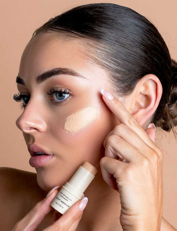 A blue-eyed woman with smooth skin applies Avocado Zinc SPF50 Natural Concealer - Light-Medium to her cheek, blending it with her finger. Her neat eyebrows and pulled-back hair stand out against a light beige background.