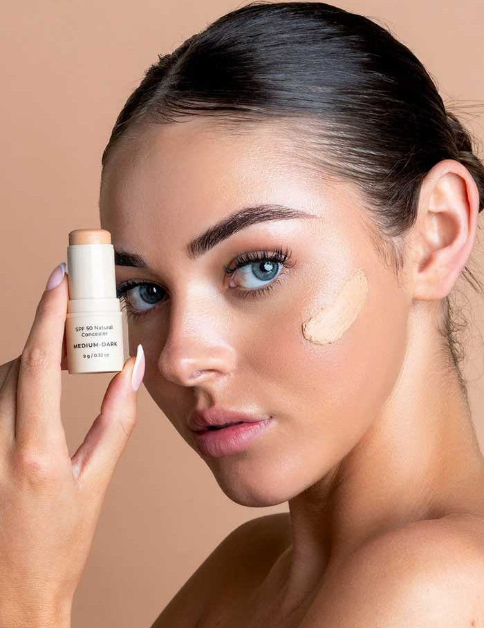 A woman with smooth skin and blue eyes holds the Avocado Zinc SPF50 Natural Concealer - Medium-Dark 9g near her face, showing a cheek swatch. She has slicked-back dark hair against a peach background.