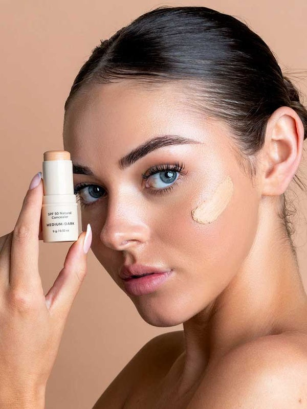 Model holding Avocado Zinc SPF50 Natural Concealer in Medium-Dark beside the cheek swatch.