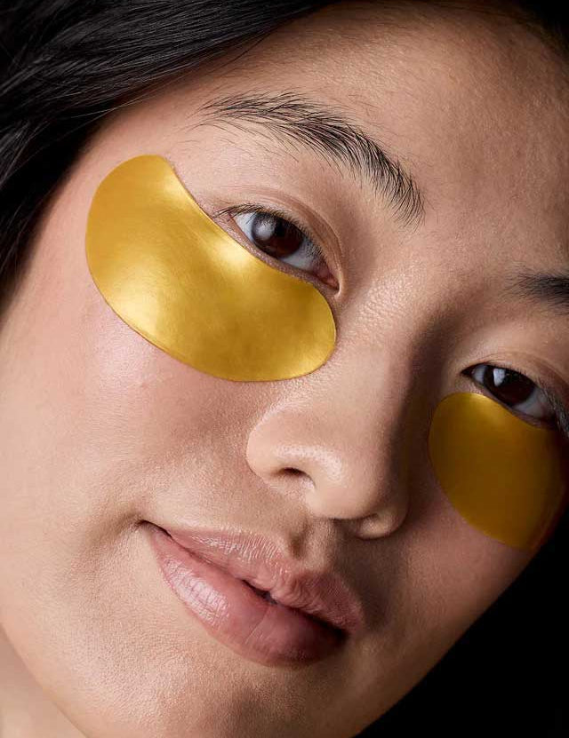 A close-up of a person with smooth skin wearing Acure Radically Rejuvenating Under Eye Hydrogels 4g gold-colored patches beneath their eyes, likely for skincare or to reduce puffiness.