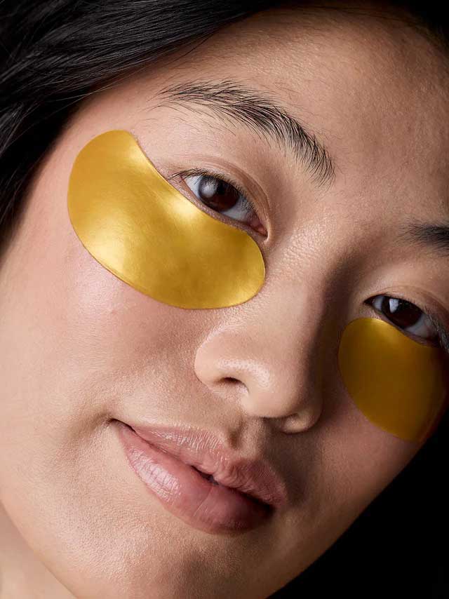 A close-up of a person with smooth skin wearing Acure Radically Rejuvenating Under Eye Hydrogels 4g gold-colored patches beneath their eyes, likely for skincare or to reduce puffiness.