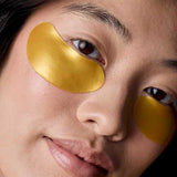 Model wearing gold Acure Radically Rejuvenating Under Eye Hydrogels beneath both eyes.