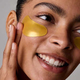 A smiling person applies Acure Radically Rejuvenating Under Eye Hydrogels 4g, gently touching their cheek. The image highlights their radiant eyes, glowing smile, and nourishing skincare routine with these under-eye patches.