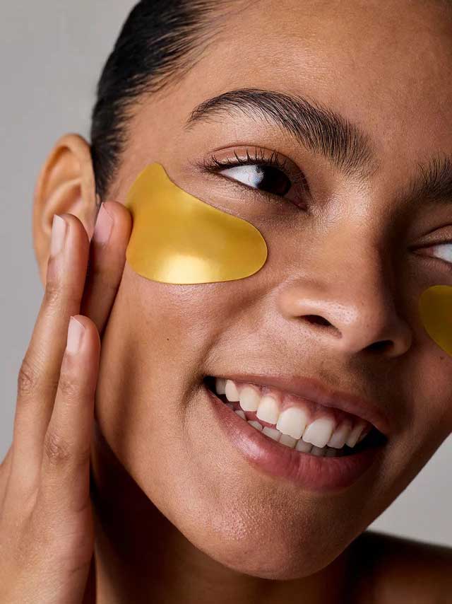 A smiling person applies Acure Radically Rejuvenating Under Eye Hydrogels 4g, gently touching their cheek. The image highlights their radiant eyes, glowing smile, and nourishing skincare routine with these under-eye patches.