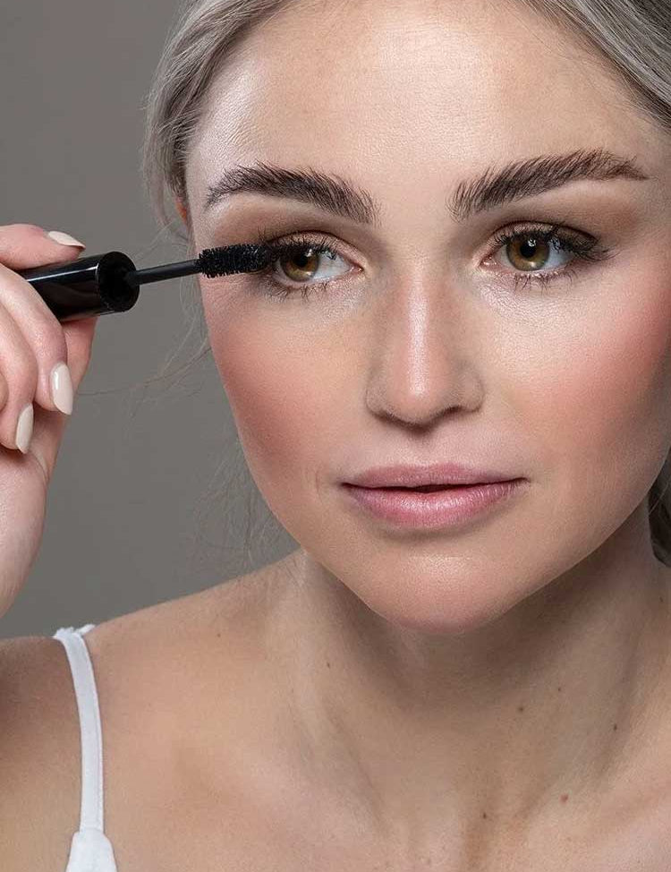A woman with light skin and blonde hair applies Benecos Mascara Maximum Volume to her upper lashes. She wears a white tank top, has natural makeup with rosy cheeks and nude lips, set against a plain neutral background.