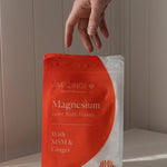 Amazing You Joint Magnesium Bath Flakes 800g orange-and-white pouch on a sunlit ledge, with a hand above it and the MSM and ginger label visible.