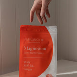 Amazing You Joint Magnesium Bath Flakes 800g orange-and-white pouch on a sunlit ledge, with a hand above it and the MSM and ginger label visible.