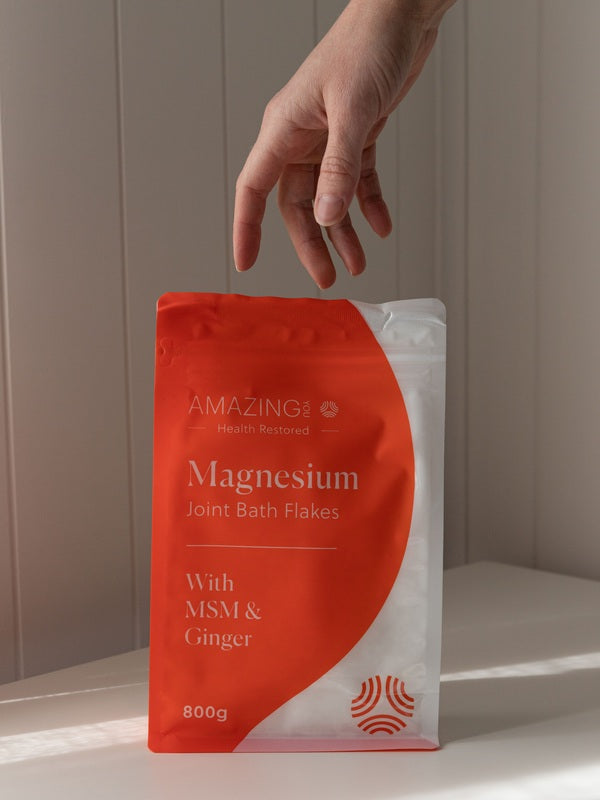 Amazing You Joint Magnesium Bath Flakes 800g orange-and-white pouch on a sunlit ledge, with a hand above it and the MSM and ginger label visible.