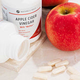 Two red apples, a bottle of Nature's Help Apple Cider with Mother Vinegar, a white bottle on its side, and several white capsules on light marble—ideal for energy boost and improved skin health.