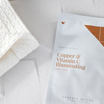 White Vanessa Megan Copper & Vitamin C Illuminating Sheet Mask sachet with copper-coloured lettering, lying beside a folded white towel on a tiled surface.