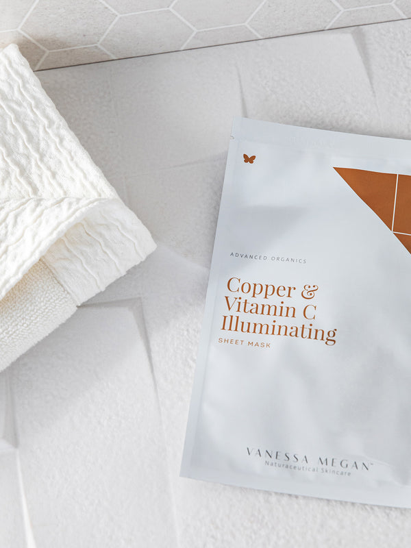 White Vanessa Megan Copper & Vitamin C Illuminating Sheet Mask sachet with copper-coloured lettering, lying beside a folded white towel on a tiled surface.