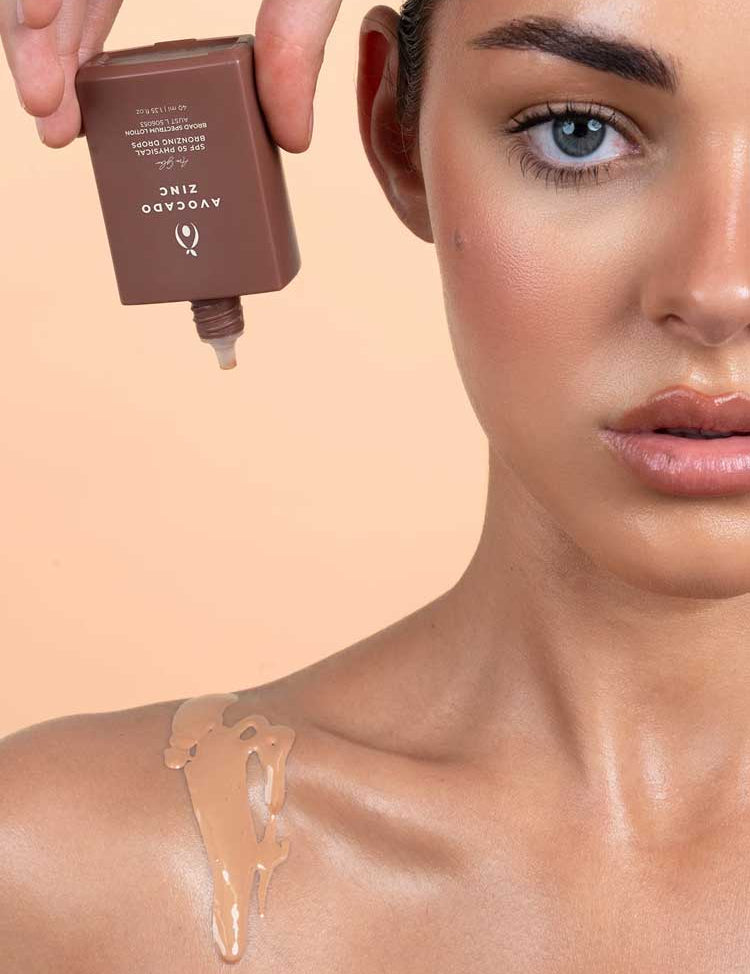 A woman with flawless skin has Avocado Zinc SPF50 Physical Bronzing Drops 40ml poured from a brown bottle onto her shoulder, showcasing smooth texture and an even, radiant complexion.